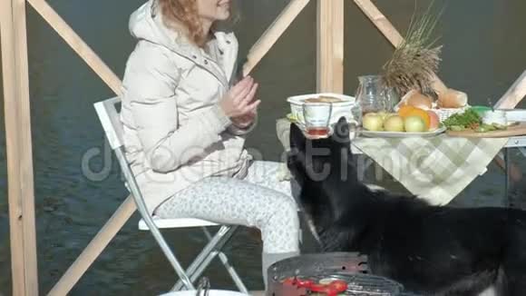戴墨镜的年轻女子穿着暖和的衣服坐在一张桌子旁喝茶吃果酱煎饼喂狗视频的预览图