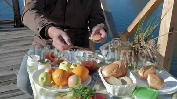 成熟的人穿着温暖的衣服把果酱放在煎饼上吃东西在木桥上河边野餐周末寒冷的天气视频的预览图