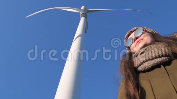 戴墨镜的年轻女性站在风车的大叶下对抗天空视频的预览图