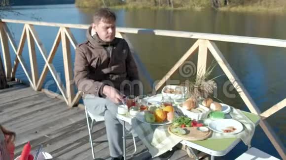 成熟的男人穿着暖和的衣服坐在桌子旁吃汉堡包加番茄酱在河岸上野餐视频的预览图