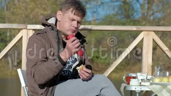 成熟的男人穿着暖和的衣服坐在桌子旁吃汉堡包加番茄酱在河岸上野餐视频的预览图