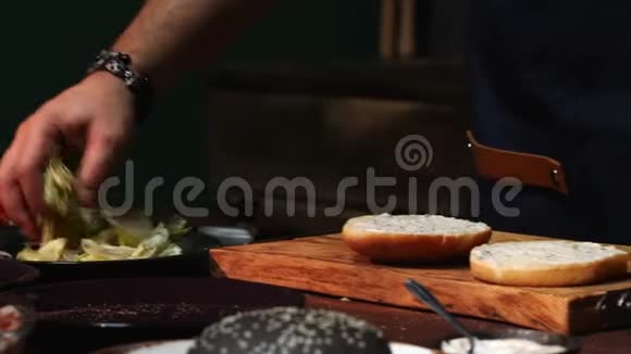 男厨师穿蓝色围裙在汉堡包上加入不同种类的绿色库存录像筹备视频的预览图