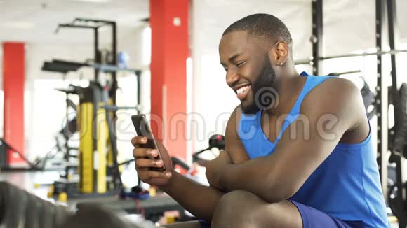 快乐的非裔美国男性坐在健身房用手机看视频视频的预览图