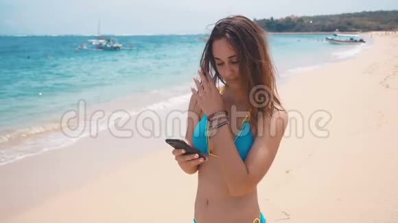年轻女性使用智能手机在海滩的海洋背景微笑的女游客在巴厘岛度假时穿着泳衣视频的预览图