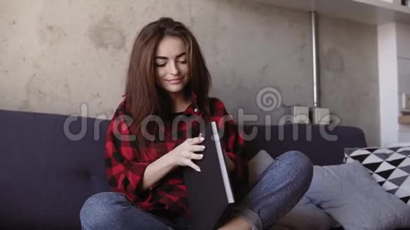 漂亮的年轻黑发女孩穿着舒适的红色法兰绒衬衫坐在沙发上看书淘汰时间视频的预览图
