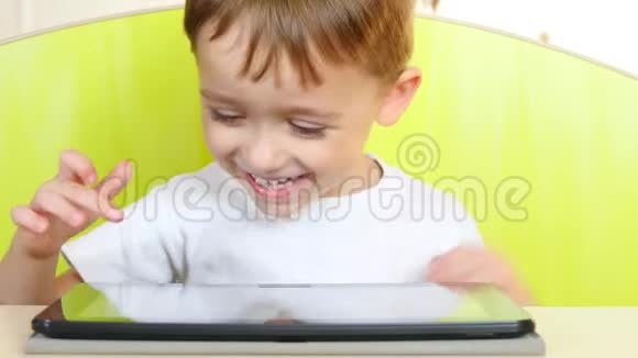 一个快乐的小男孩坐在桌子旁触摸平板电脑的电子显示器一个孩子在玩手提电脑视频的预览图