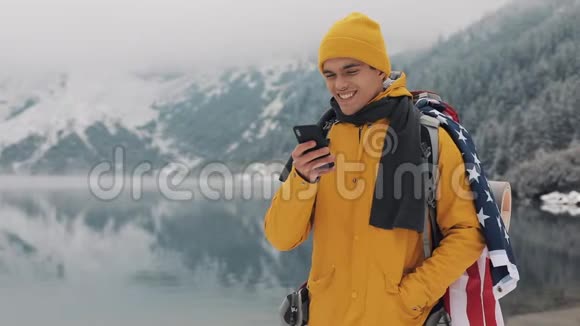 美国旅行者在徒步冬季旅行中使用智能手机白雪皑皑的湖面和背景下的群山旅行和视频的预览图