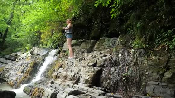 女孩博客作者正在领导一份报告坐在瀑布旁的岩石上美丽的瀑布和美丽的自然景观视频的预览图