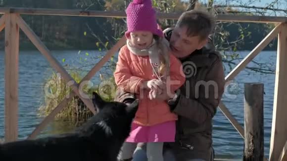 爸爸和女儿成熟的男人和小女孩穿着温暖的衣服微笑着喂狗在河岸上的木制野餐视频的预览图