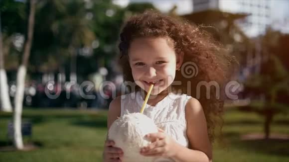 小白种人卷发的小女孩穿着白色连衣裙喝着椰子视频的预览图
