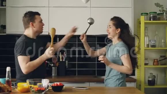 快乐的一对夫妇在厨房里用大勺子击剑在家做早餐时互相拥抱视频的预览图
