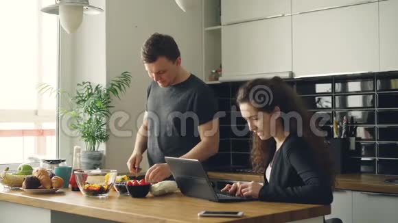 年轻漂亮的白种人女商人在家里用笔记本电脑工作丈夫正站在桌子旁边切东西视频的预览图