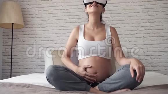 美丽的年轻孕妇在家中坐在床上时使用虚拟现实眼镜视频的预览图