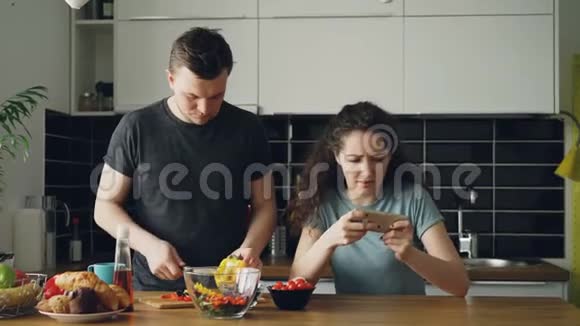 家里厨房里的漂亮情侣可爱的女孩在智能手机上玩电子游戏而她的男朋友在做饭视频的预览图