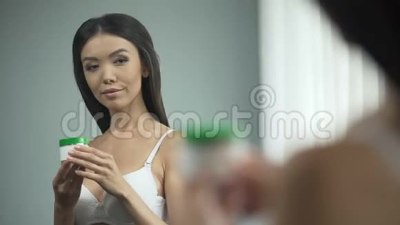 美丽的亚洲女孩脸上涂着奶油站在镜子前美丽视频的预览图