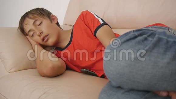 年轻人睡在沙发上白天穿红色t恤和蓝色牛仔裤睡在米色皮革沙发上的白人少年视频的预览图