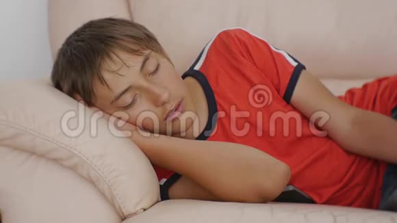 年轻人睡在沙发上白天穿红色t恤和蓝色牛仔裤睡在米色皮革沙发上的白人少年视频的预览图