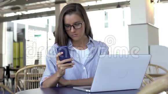 在笔记本电脑上工作时戴眼镜的女人用智能手机发短信视频的预览图