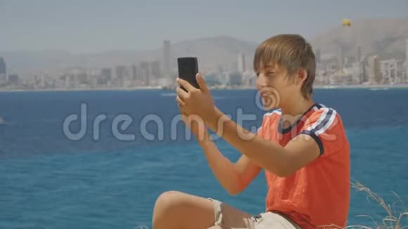 穿着红色t恤和短裤的英俊少年在海上城市天际线上播放智能手机视频视频的预览图