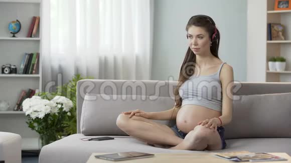未来的母亲坐在沙发上戴着耳机伸展背部进行音频锻炼视频的预览图