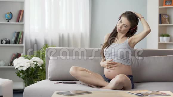 年轻孕妇戴着耳机坐在沙发上做伸展运动视频的预览图