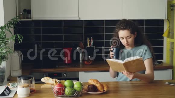 漂亮的卷发女人坐在桌子旁端着杯咖啡在家里漂亮的厨房里喝酒看书她是视频的预览图