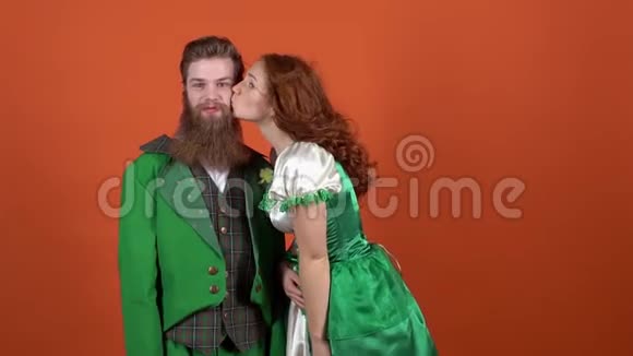 穿着圣诞服装的年轻夫妇庆祝圣帕特里克节橙色墙上的妻子亲吻丈夫视频的预览图