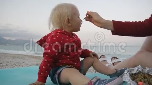 金发女娃在海边的背景下妈妈手里拿着食物吃勺子玩得很开心孩子和妈妈视频的预览图