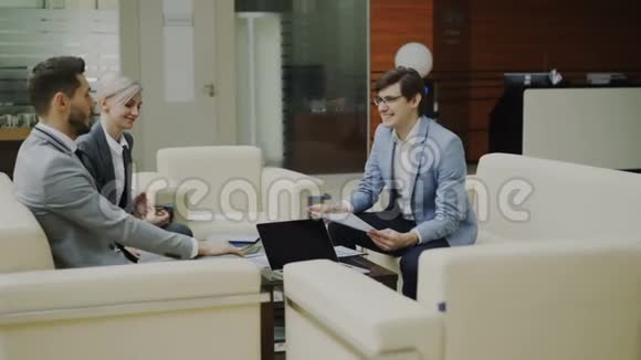 快乐的商人和商业伙伴坐在现代办公大厅的沙发上讨论财务报告视频的预览图