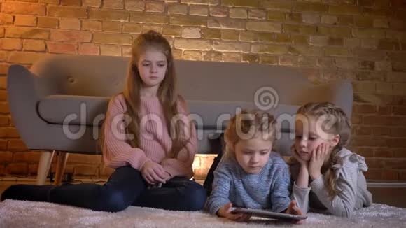 三个白种人小女孩坐在地板上观看平板电脑的肖像她的注意力和兴趣都集中在视频的预览图