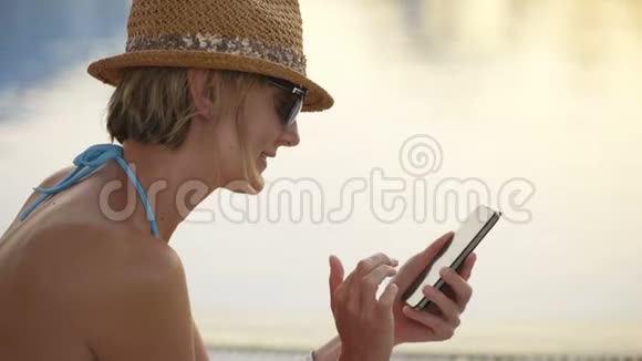 穿泳装的女人坐在游泳池边玩智能手机视频的预览图