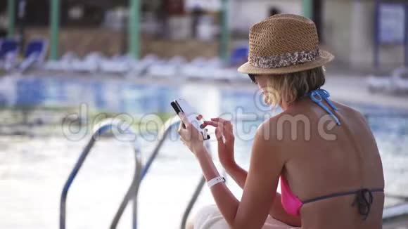 穿泳装的女人坐在游泳池边玩智能手机视频的预览图