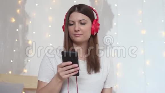 美丽的女孩听音乐耳机红色坐在床上放松使用手机家风视频的预览图