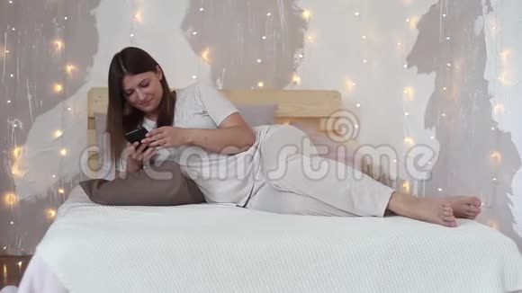 穿着睡衣的快乐漂亮女孩喜欢手机躺在家里的床上视频的预览图