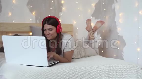 美丽快乐的女孩躺在一张大床上用一台白色的移动电脑用耳机听音乐视频的预览图