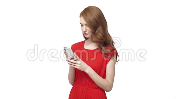 漂亮的年轻女子穿着橙色连衣裙一边用手机发短信给朋友在白色背景上孤立视频的预览图