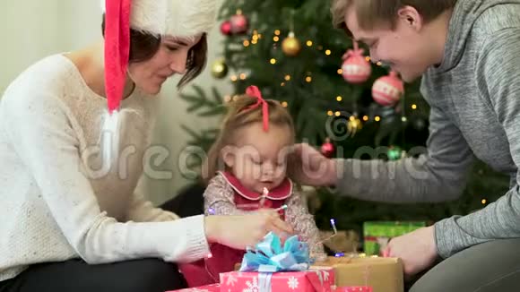 幸福的家庭父母在圣诞树前向他们可爱的孩子展示新年装饰品妈妈戴着圣诞帽视频的预览图