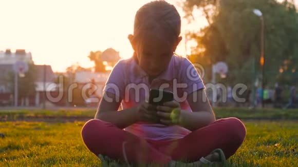 可爱的女孩用智能手机坐在中央公园的草坪上年轻貌美的女孩在手机上做手势视频的预览图