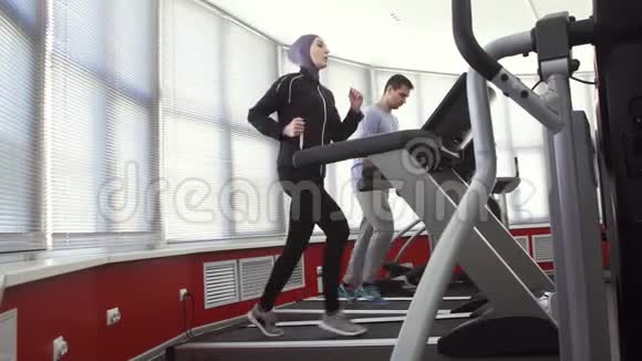 女运动员戴头巾在跑步模拟器和一个男人在健身房家庭视频的预览图