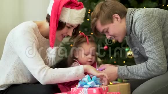 幸福的家庭父母在圣诞树前向他们可爱的孩子展示新年装饰品妈妈戴着圣诞帽视频的预览图