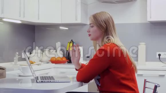 漂亮的金发美女坐在厨房里用电玩与朋友交谈视频的预览图