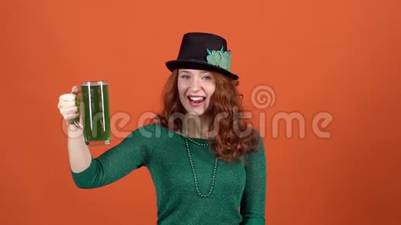 年轻女子庆祝圣帕特里克节橙色墙上戴着帽子竖起大拇指喝绿色啤酒视频的预览图