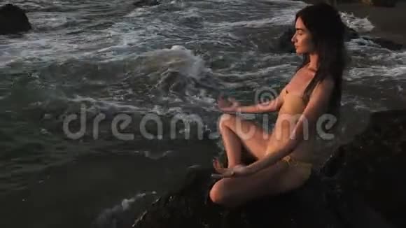 女孩坐在海边的岩石上沉思着莲花瑜伽的姿势全神贯注视频的预览图