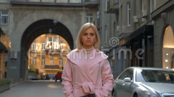 一个年轻漂亮的金发女郎穿着粉红色的外套站在现代城市的街道上视频的预览图