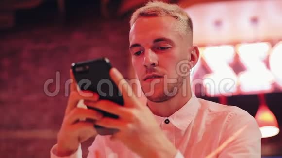 晚上使用智能手机的年轻魅力男子他坐在靠近霓虹灯标志的酒吧或餐馆里通信视频的预览图