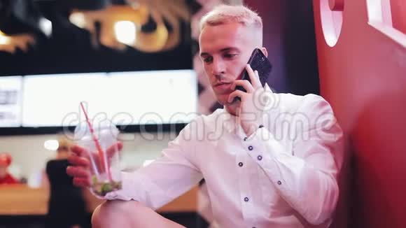 年轻的帅哥在夜间咖啡馆的智能手机上聊天他坐在霓虹灯招牌旁边喝着鸡尾酒视频的预览图