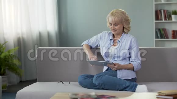 快乐的中年妇女在平板电脑上观看有趣的视频坐在家里的沙发上视频的预览图