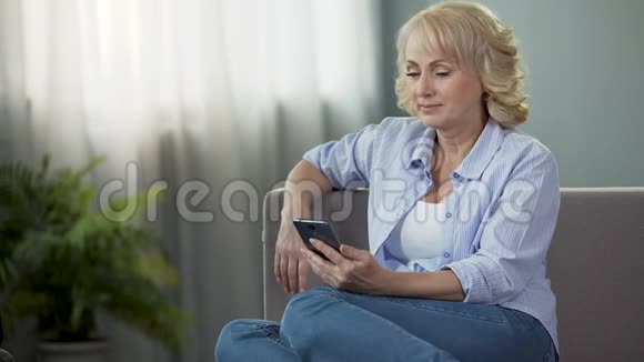 快乐的中年女性坐在沙发上通过电话观看有趣的照片视频的预览图