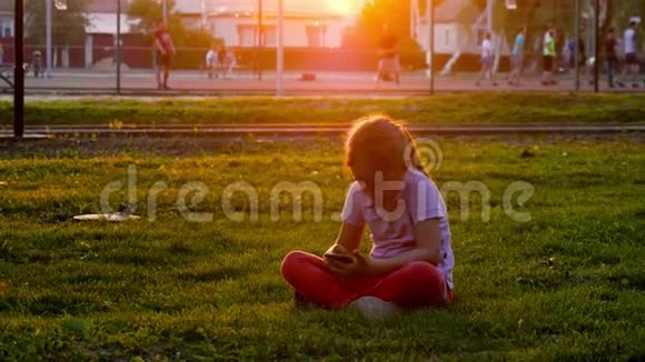 可爱的女孩用智能手机坐在中央公园的草坪上年轻貌美的女孩在手机上做手势视频的预览图