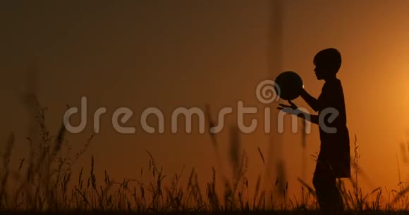 剪影一个男孩在日落时在田里戏耍一个球视频的预览图